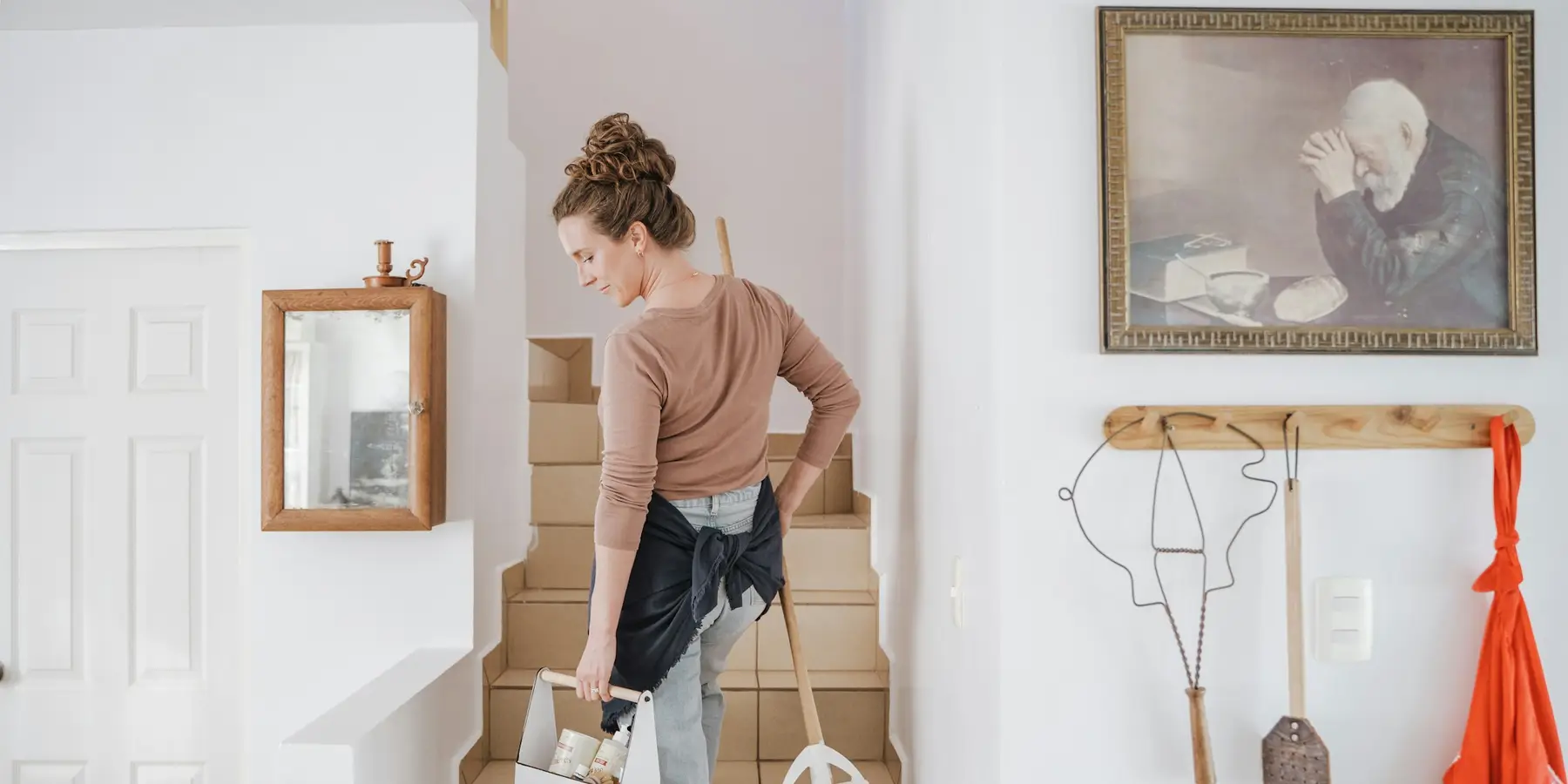 A cleaner carrying a caddy and broom up the stairs of a home